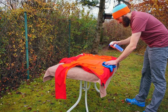 Mann spr&uuml;ht rot-orangene Regenjacke mit Impr&auml;gniermittel ein.