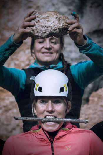 Eine Frau mit Kletterhelm &uuml;ber dem eine andere Frau bedrohlich einen Felsbrocken h&auml;lt.