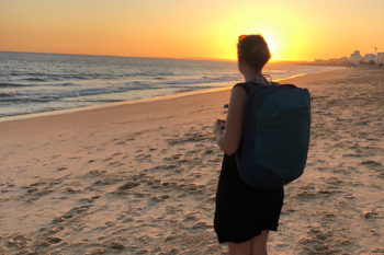 Eine Frau steht mit Rucksack am Strand und blickt in den Sonnenuntergang.