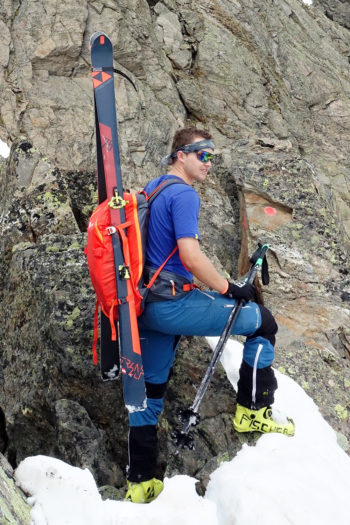 Ein Skibergsteiger steht am Fels mit dem orangen Salewa Randonnée Rucksack und Ski am Rücken.