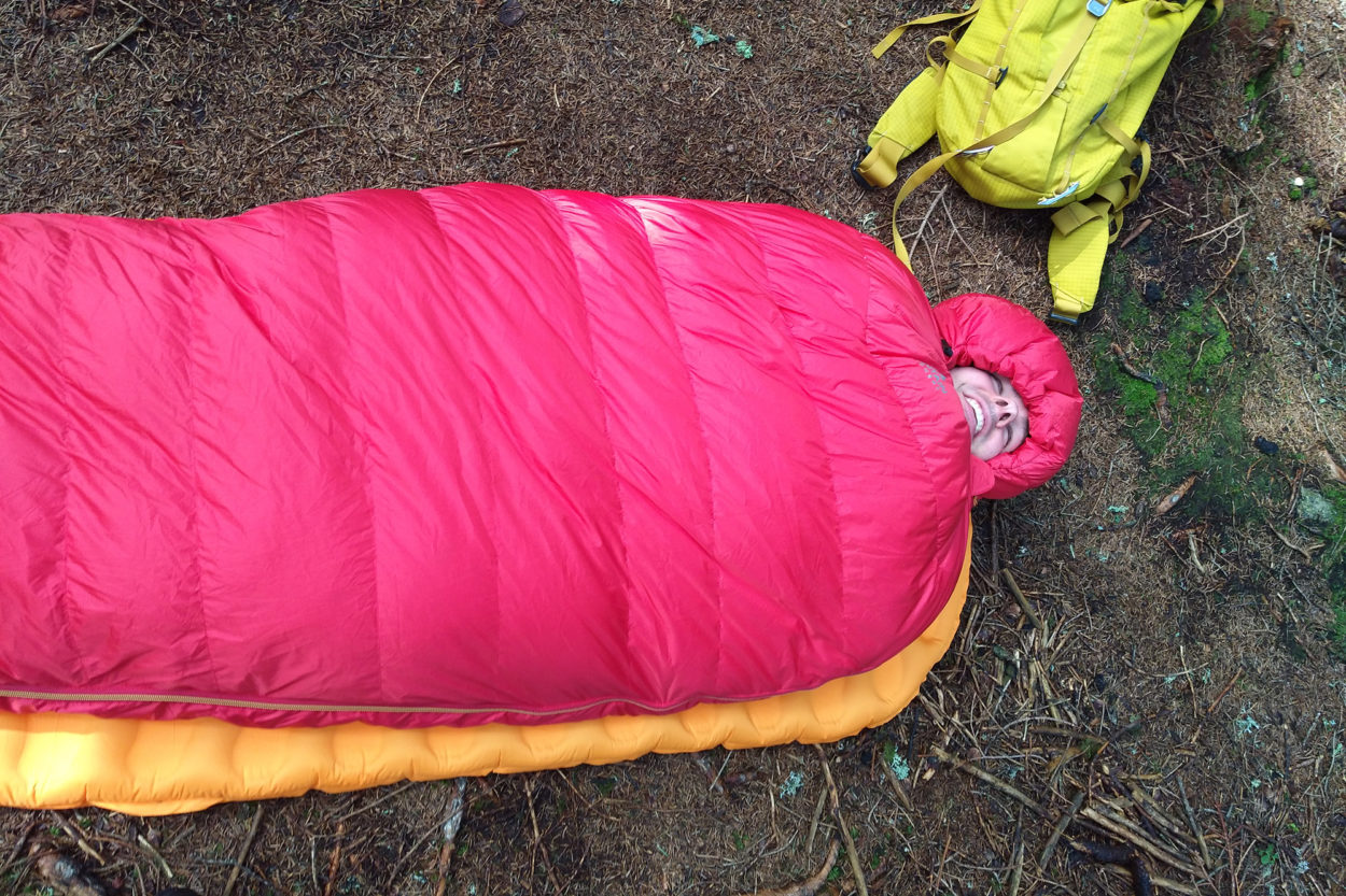 Mann liegt in rotem Schlafsack Glacier 700 auf Waldboden