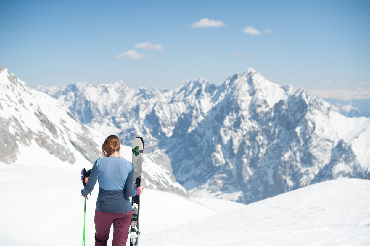 Frau steht mit Rücken zur Kamera vor winterlichem Bergpanorama und hält Ski in der Hand