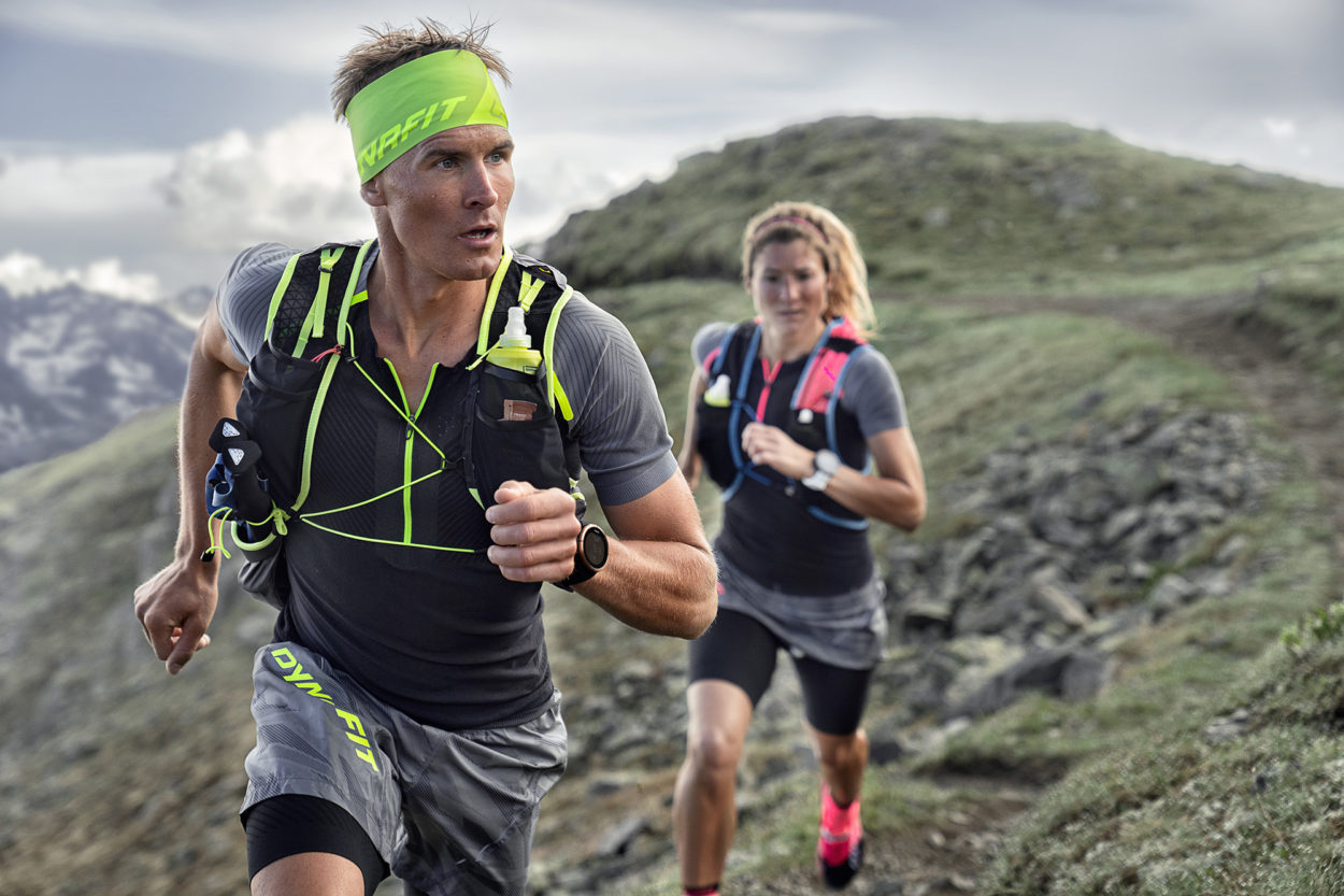 Mann im schwarz-grauen Trailrunning-Outfit, hinter ihm eine Frau in schwarz-pinken Outfit beim Trailrunning oben in den Bergen.